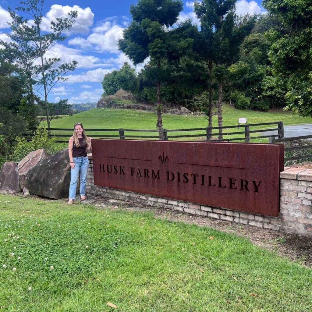 Woman smiling at camera next to a sign that reads Husk Farm Distillery.