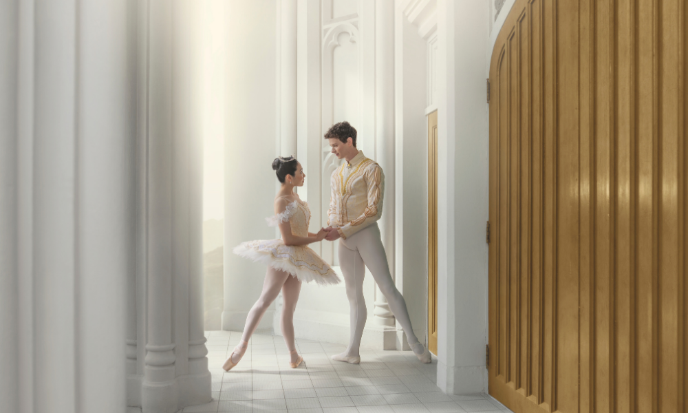 Two ballet dancers stand holding hands. 